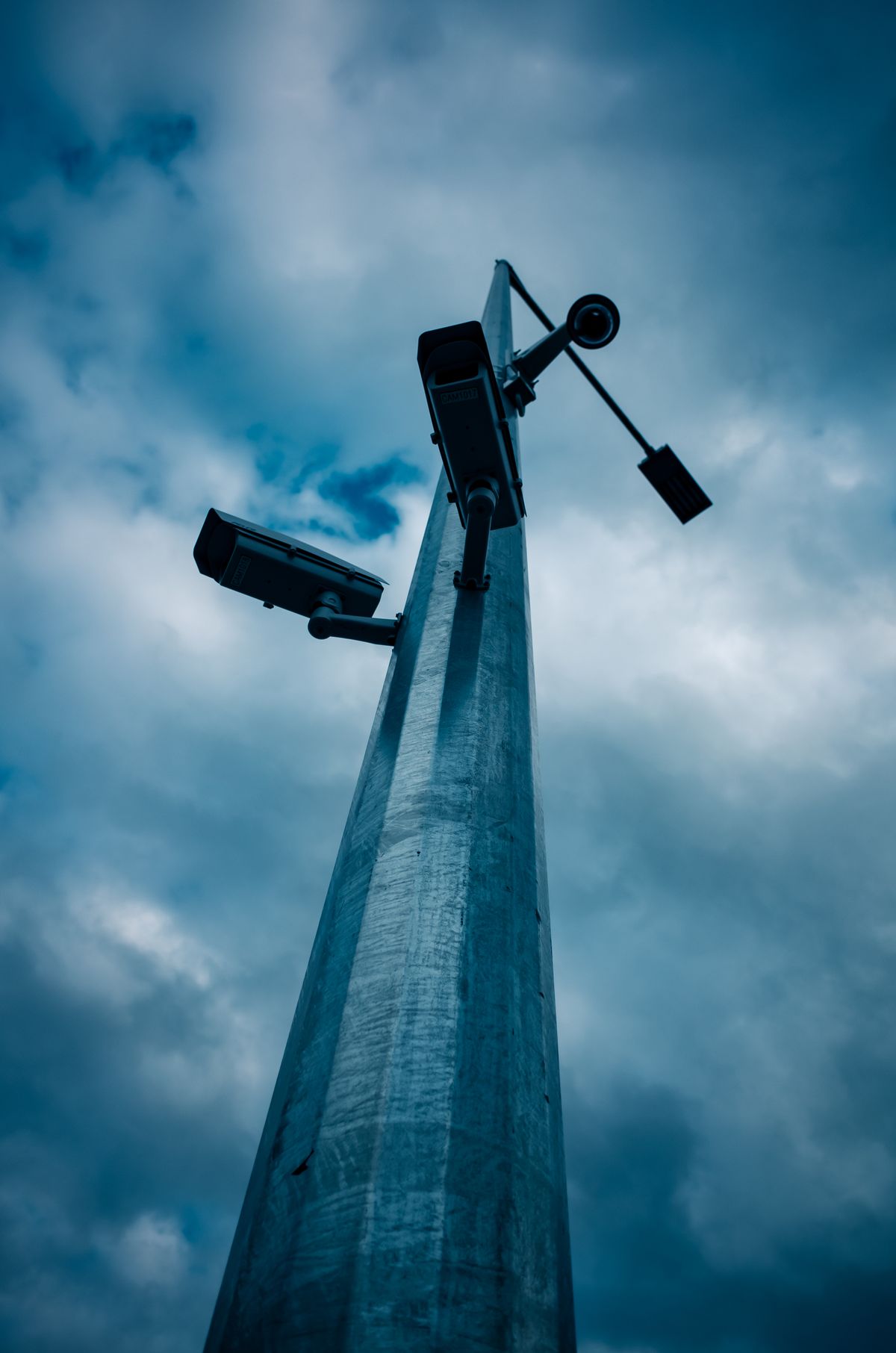 Surveillance cameras on a pole against a dark sky — the watcher knows it is being watched
