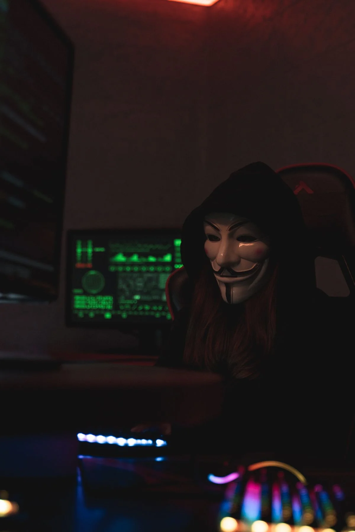 A masked hacker at a workstation with glowing green code monitors